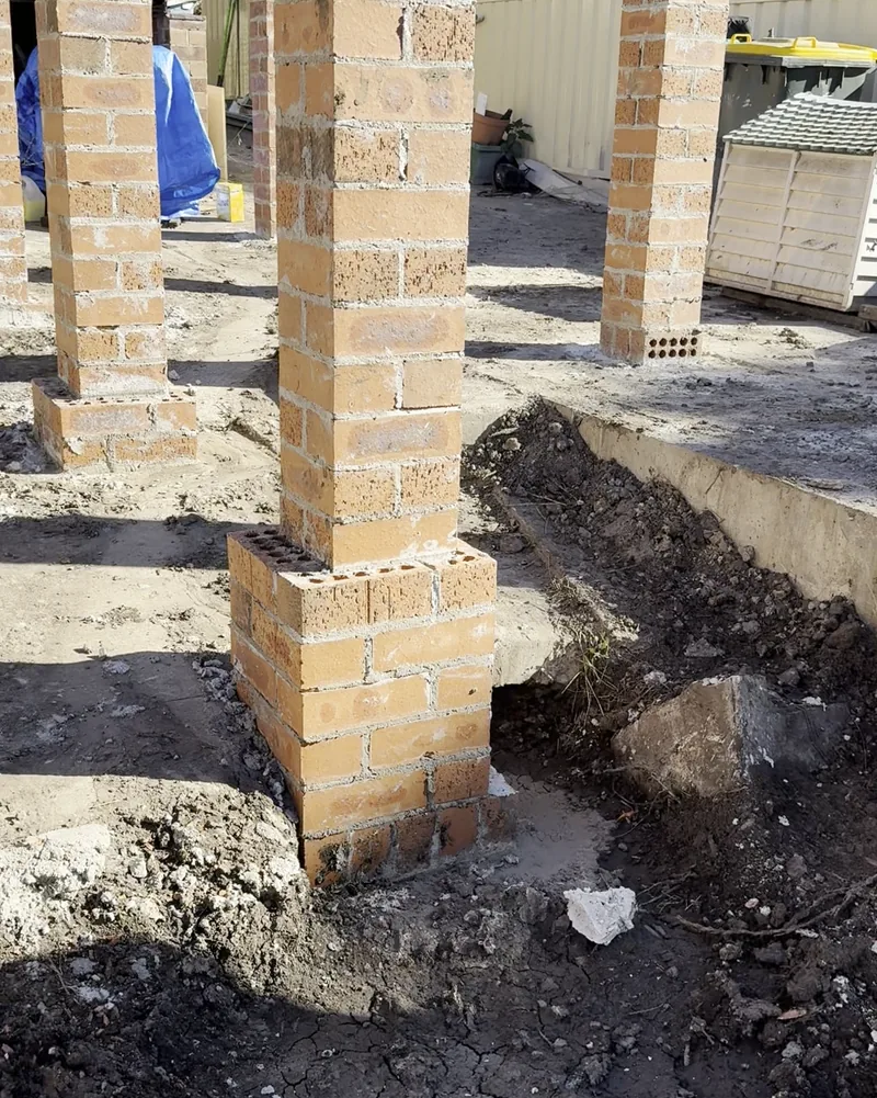 Brick piers standing on new concrete footings in an excavated area beside a house