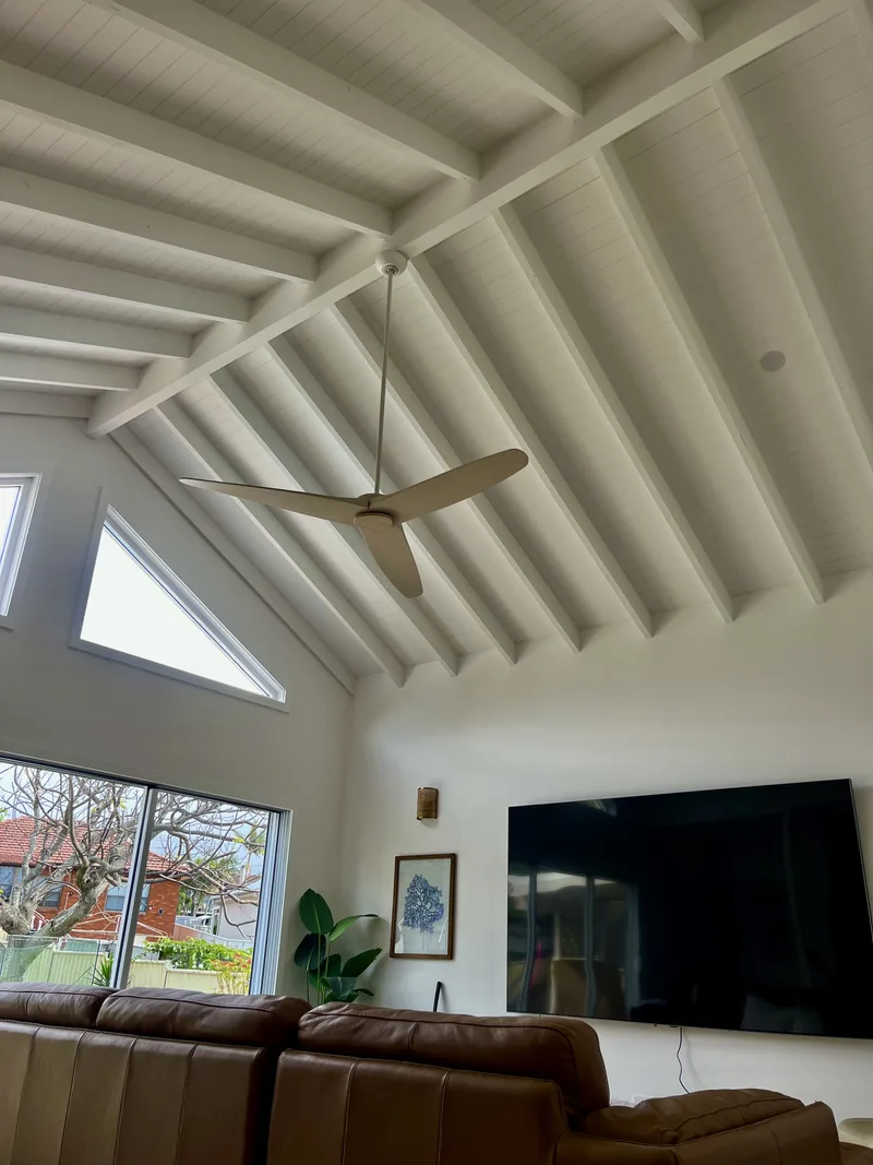 Living room with a raked ceiling, white painted rafters, a ceiling fan and a triangular high window
