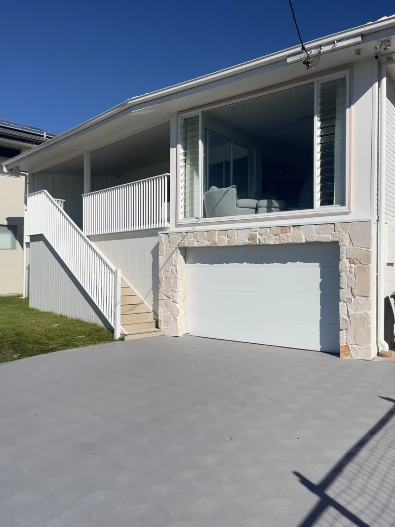 The house after the renovation: a white exterior, a covered verandah with white balustrades, new stairs, stone around a white garage door and a paved driveway