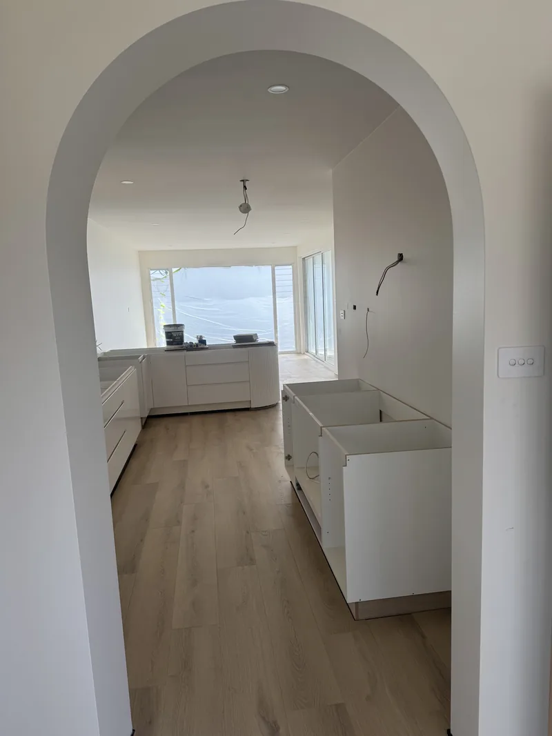 View through a curved archway into the kitchen area during fit-out, with cabinets in place and windows at the far end