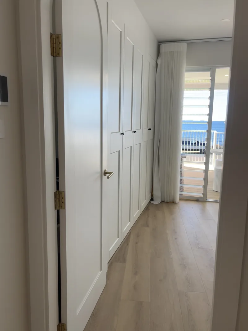 Hallway lined with white shaker-style built-in robes and timber-look flooring, leading to a balcony with an ocean view