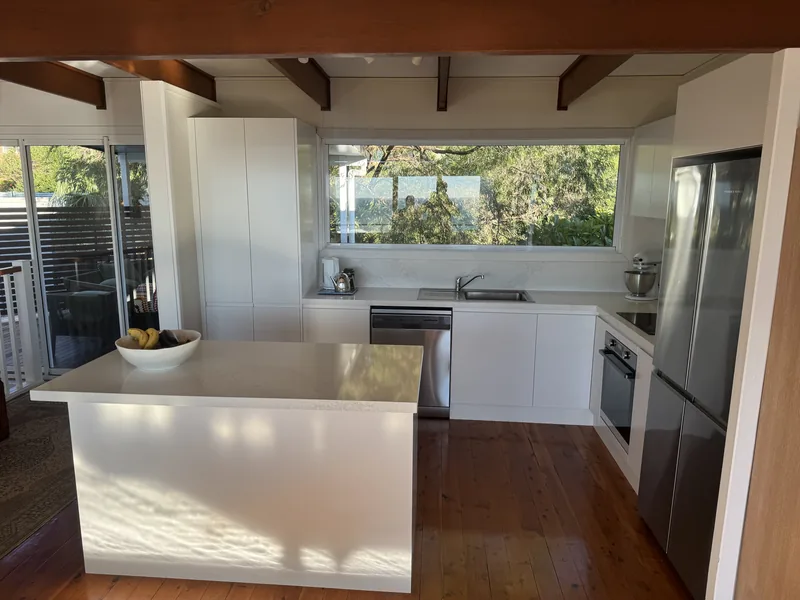 White kitchen with an island bench under exposed timber ceiling beams, a window over the sink and a stainless steel fridge