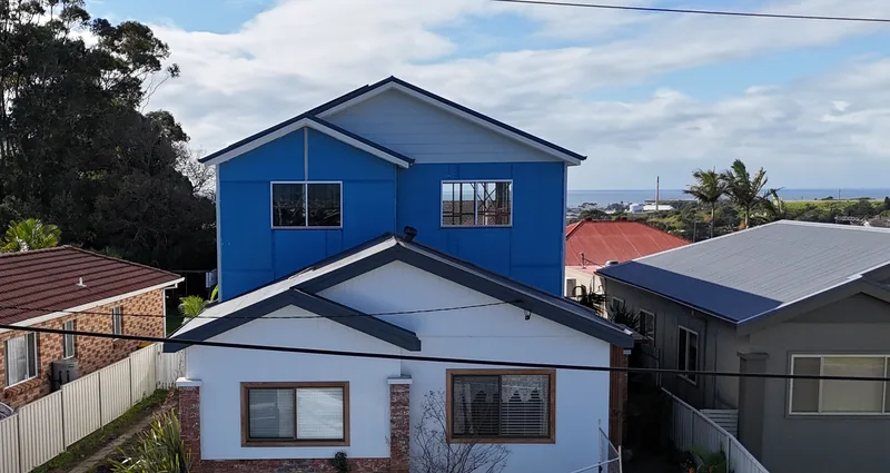 A single-storey white house with a brick base and a new two-window upper level above it, still in blue sheeting, with the ocean in the distance